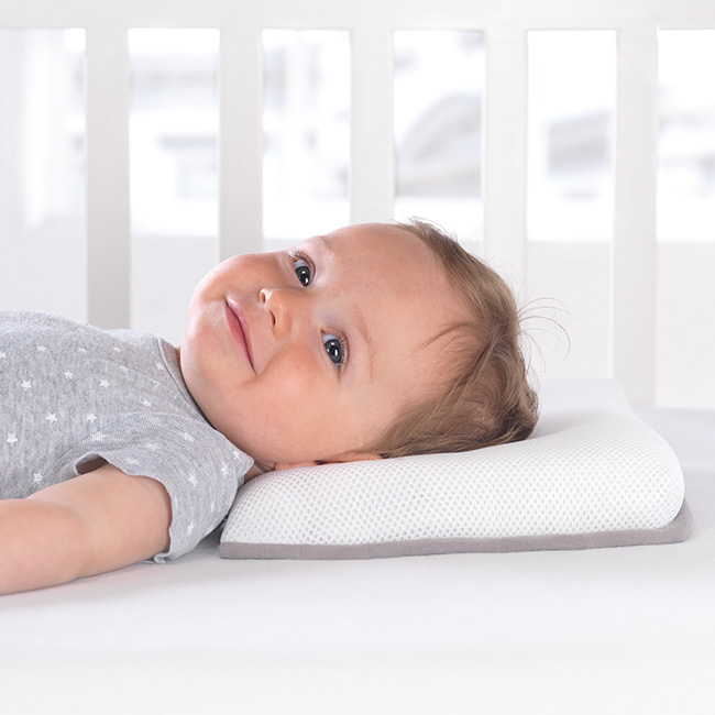 Ein lächelndes Baby liegt auf einem weißen Kissen in einem Gitterbett, trägt ein graues Shirt mit Sternenmuster, umgeben von einer ruhigen, lichtdurchfluteten Umgebung.