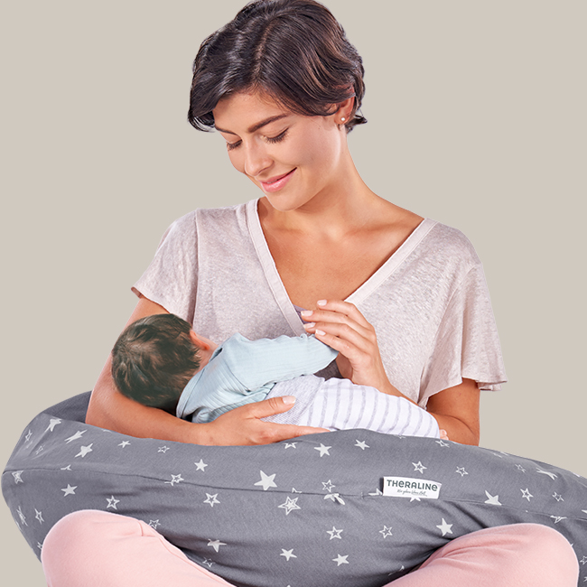 Eine Frau hält ein Baby, das eine blaue Strampler trägt, auf einem grauen Theraline-Kissen mit weißen Sternen. Sie lächelt liebevoll. Der Hintergrund ist einfarbig beige.
