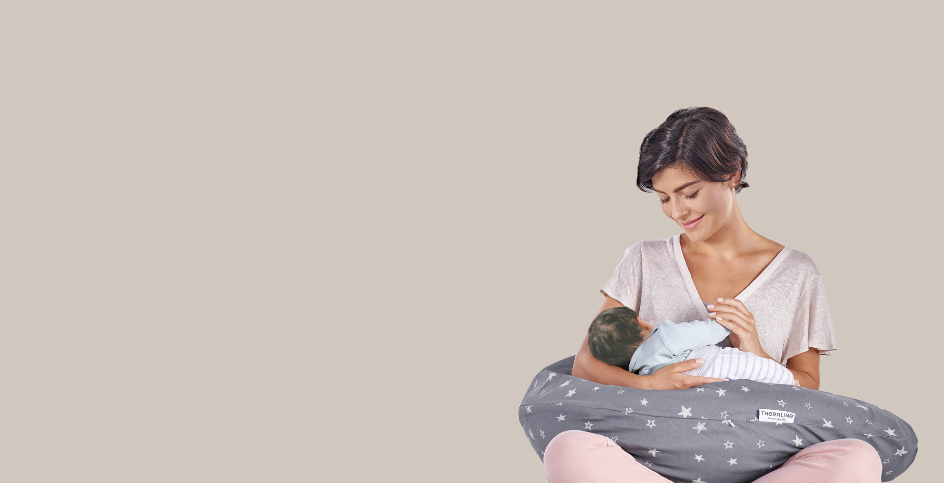 Eine Frau hält ein Baby, das von einem grauen Kissen mit weißen Sternen gestützt wird. Das Theraline-Kissen bietet Unterstützung. Der Hintergrund ist einfarbig beige.