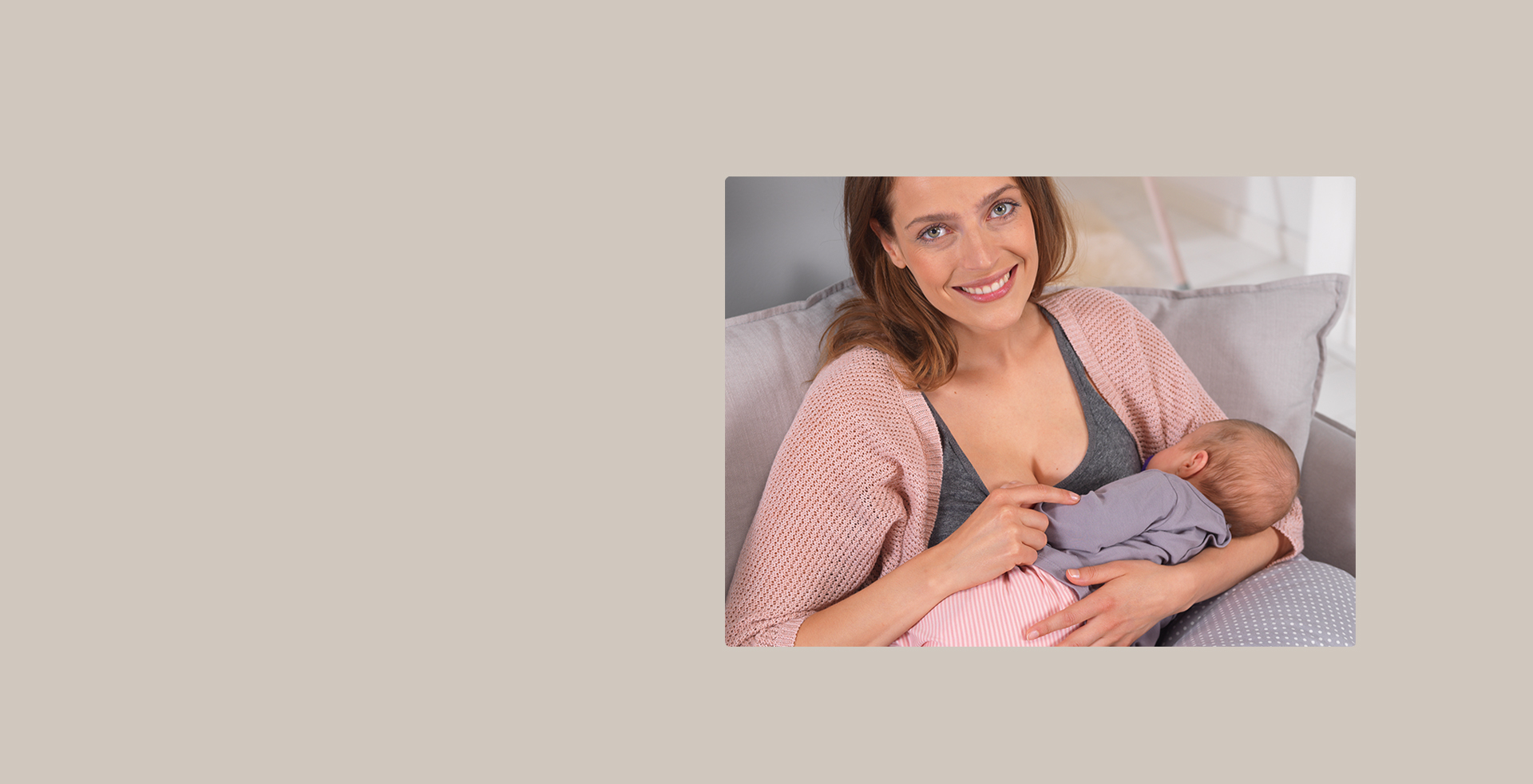 Eine Frau stillt ein Baby, während sie auf einem Sofa mit grauen Kissen sitzt. Sie trägt eine rosa Strickjacke und lächelt in gemütlicher Umgebung. Der Hintergrund ist einfarbig beige.