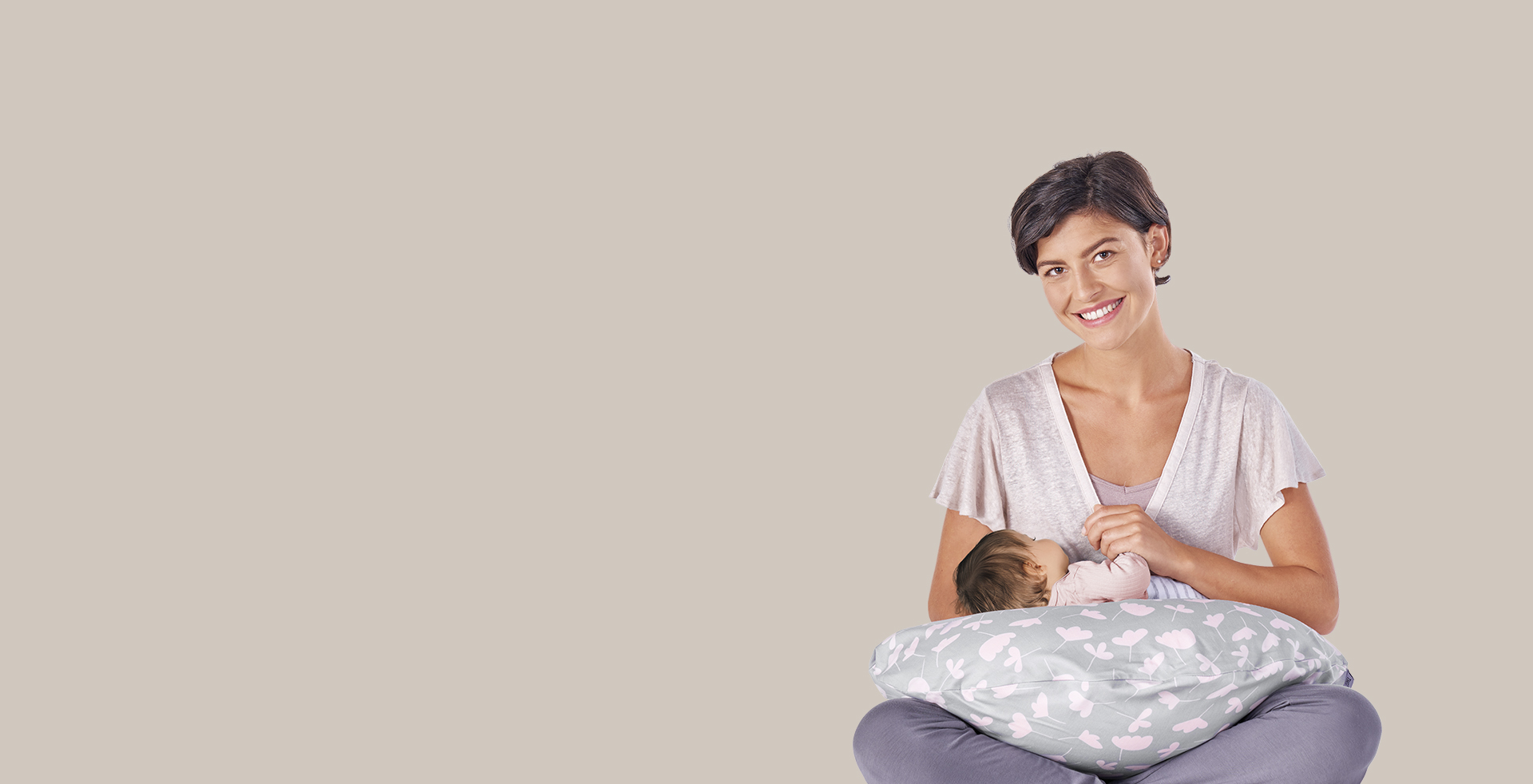 Eine Frau hält ein Baby, das auf einem Theraline-Kissen liegt. Sie sitzt lächelnd vor einem beigen Hintergrund, während das Baby mit dem Gesicht nach unten ruht.