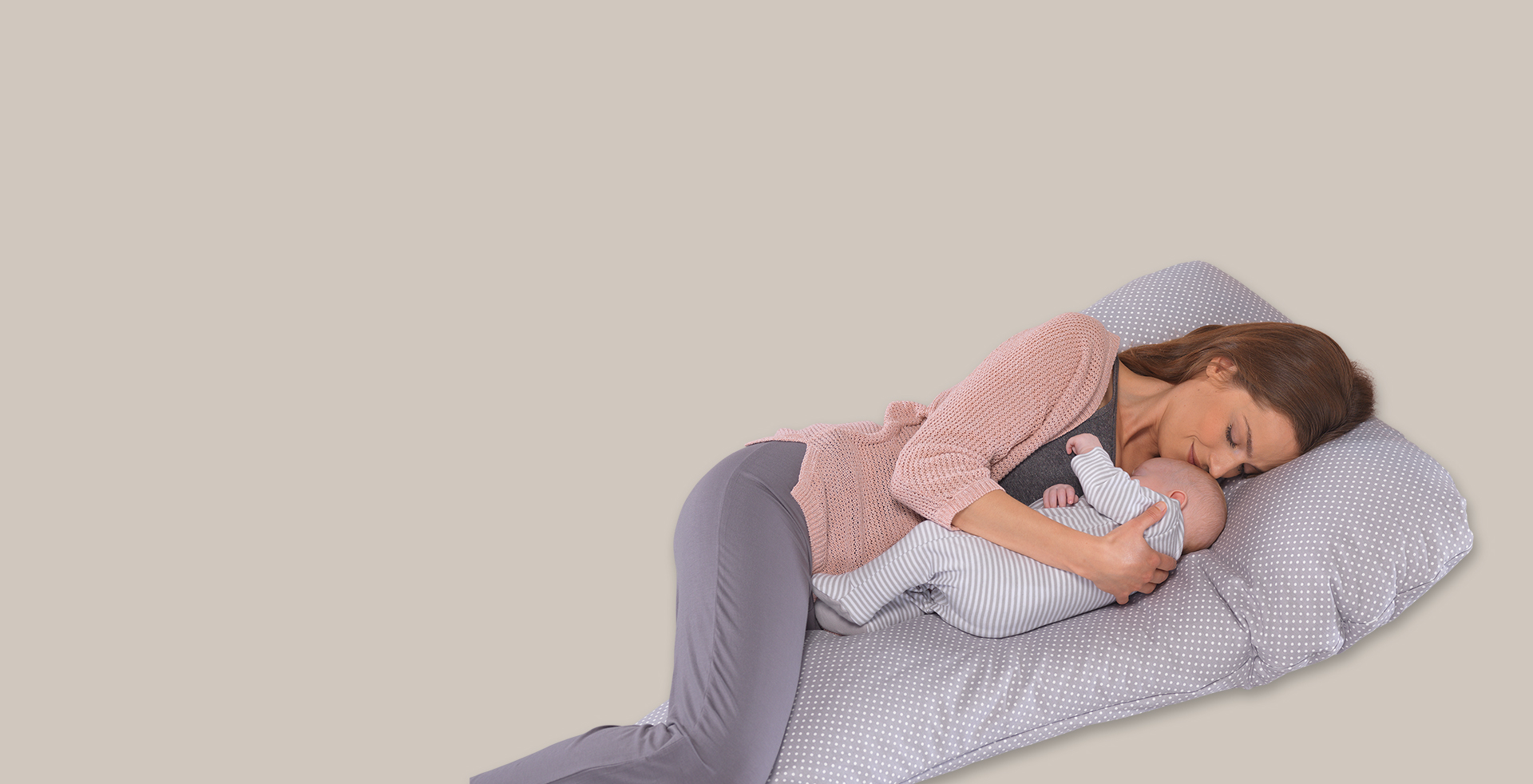 Eine Frau liegt auf einem großen, gepunkteten Kissen und hält ein Baby im Arm. Sie trägt eine rosa Strickjacke. Der Hintergrund ist einfarbig beige.