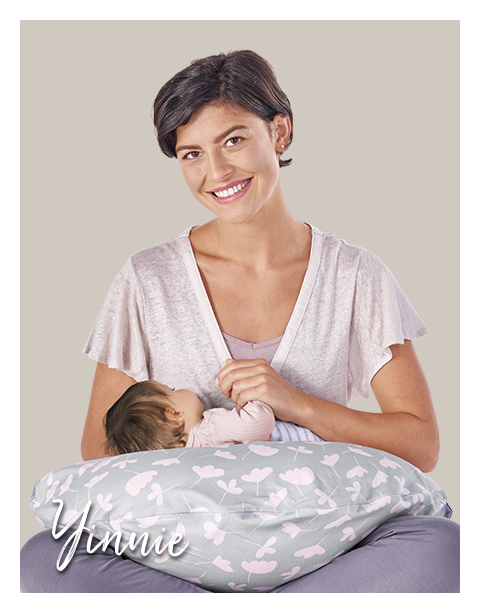 Eine Frau hält ein Baby, das auf einem Theraline Stillkissen liegt. Das Kissen hat ein Design mit weißen und rosa Blättern. Der Hintergrund ist einfarbig beige. Text: \